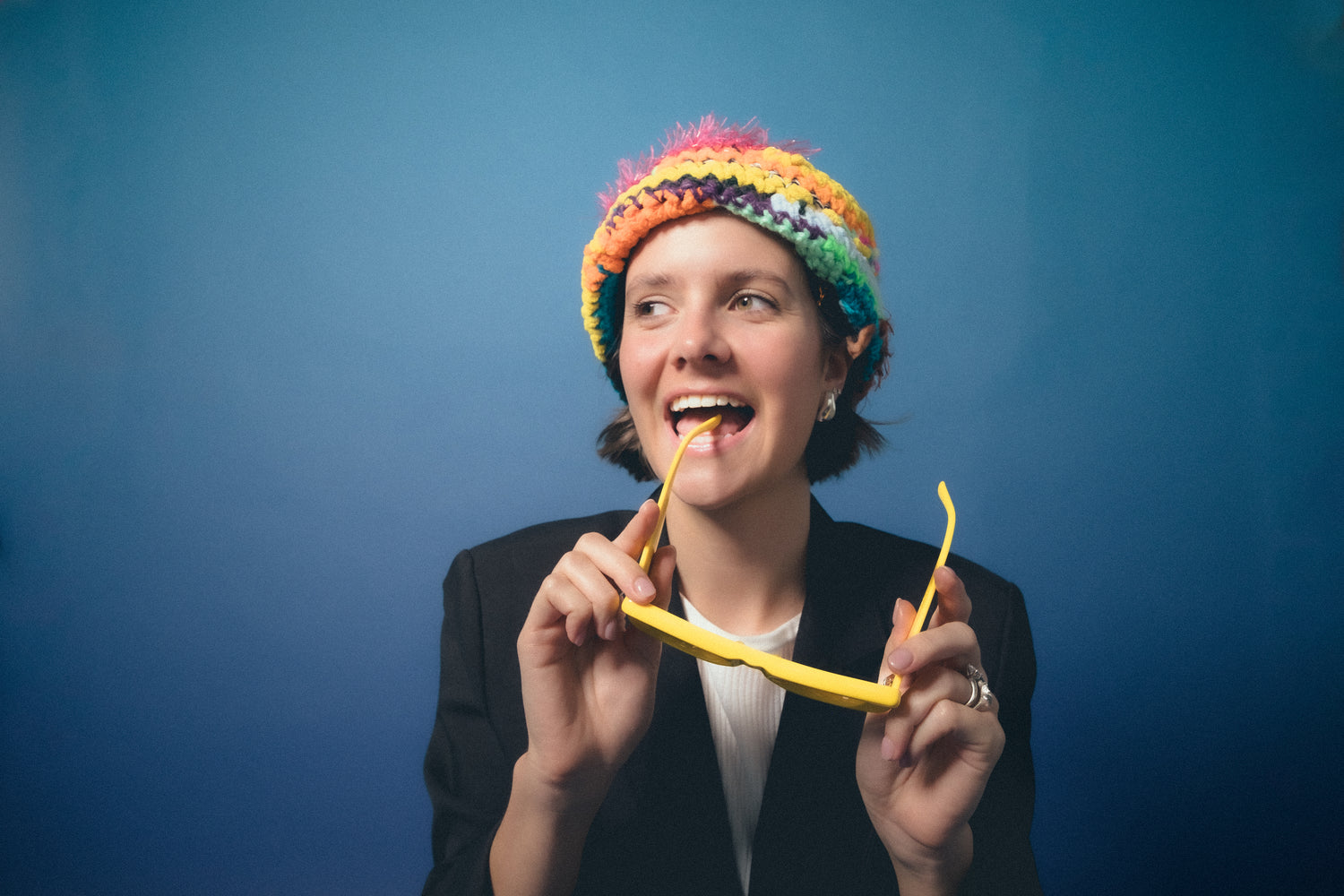 woman with short hair wearing a colorful knit beanie, white shirt, and black blazer, while holding yellow sunglasses