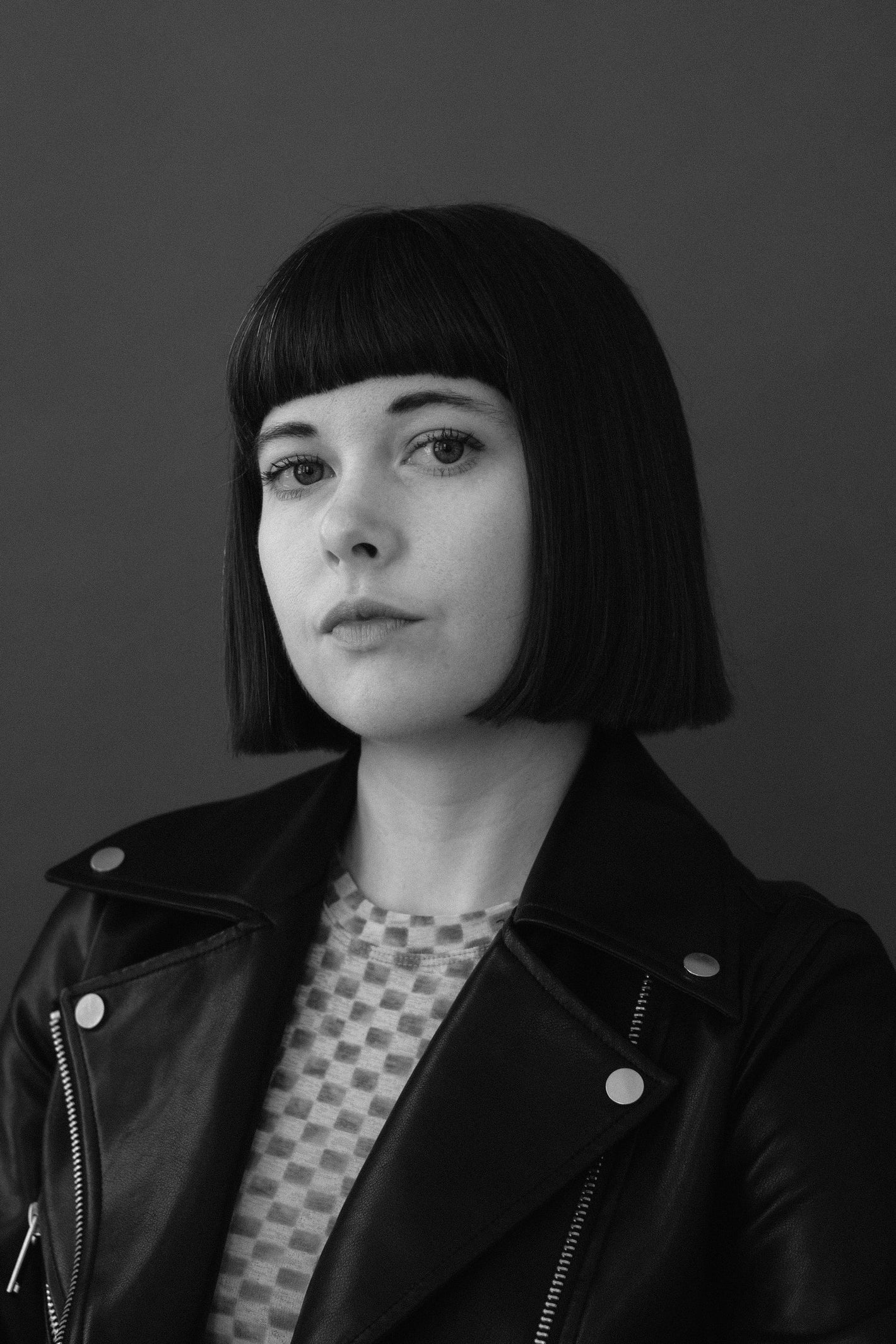 headshot of a woman with short hair and bangs wearing a checkered shirt and a leather jacket