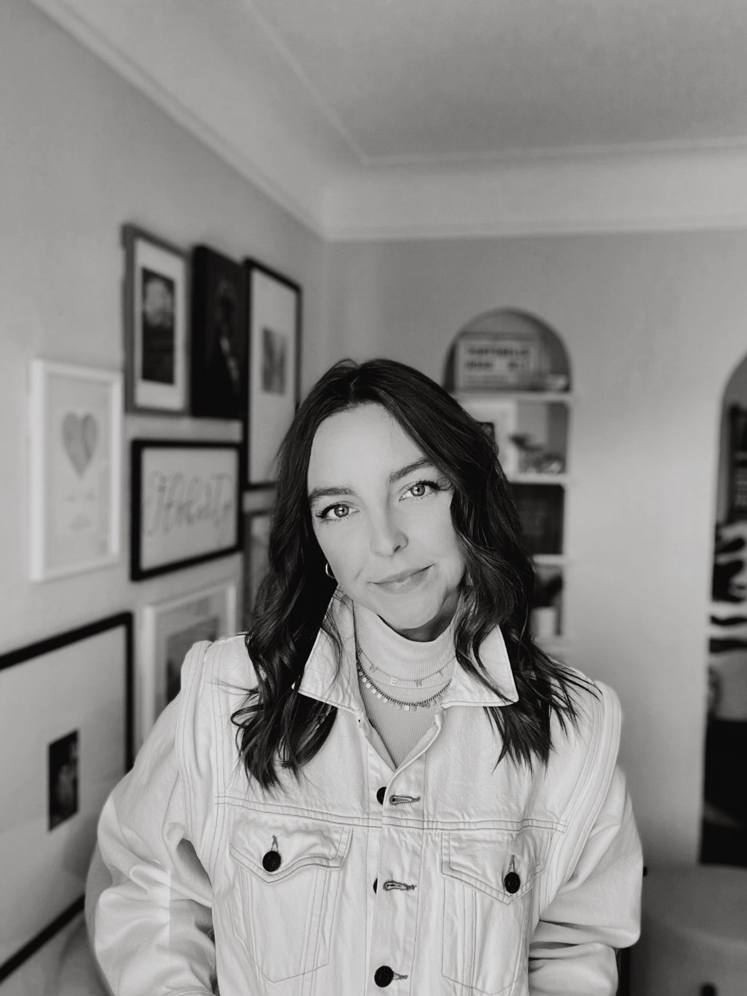 woman standing in front of a wall covered in picture frames wearing a turtleneck and jean jacket