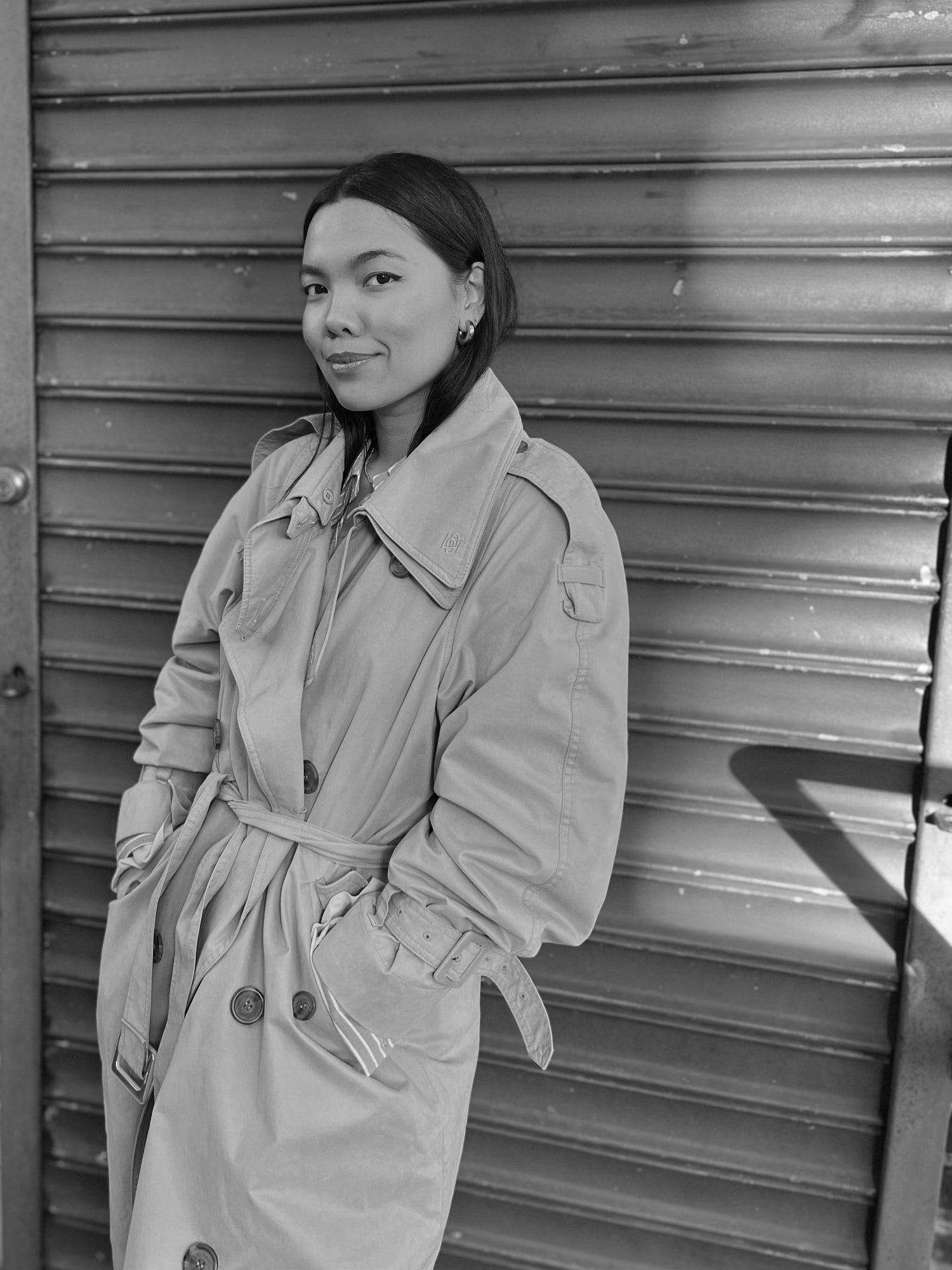 woman wearing a trench coat with her hands in her pockets smiling at the camera while leaning against a metal door